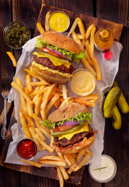 Two Brooklyn Brisket burgers with melted cheese, served with French fries, ketchup, mustard, and pickles on a wooden cutting board.